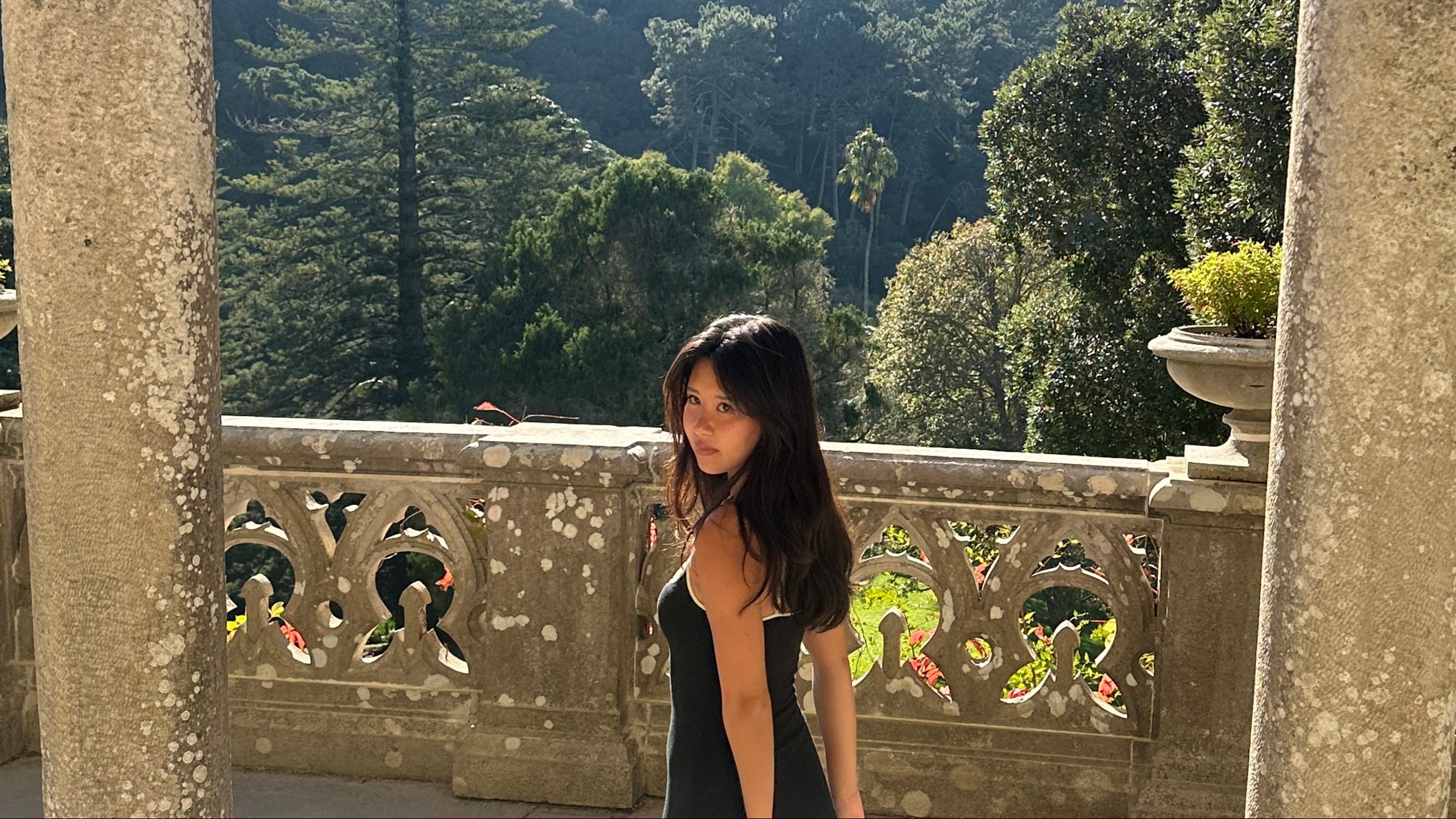 A woman wearing a black dress stands on a stone terrace with a scenic forested mountain landscape in the background.